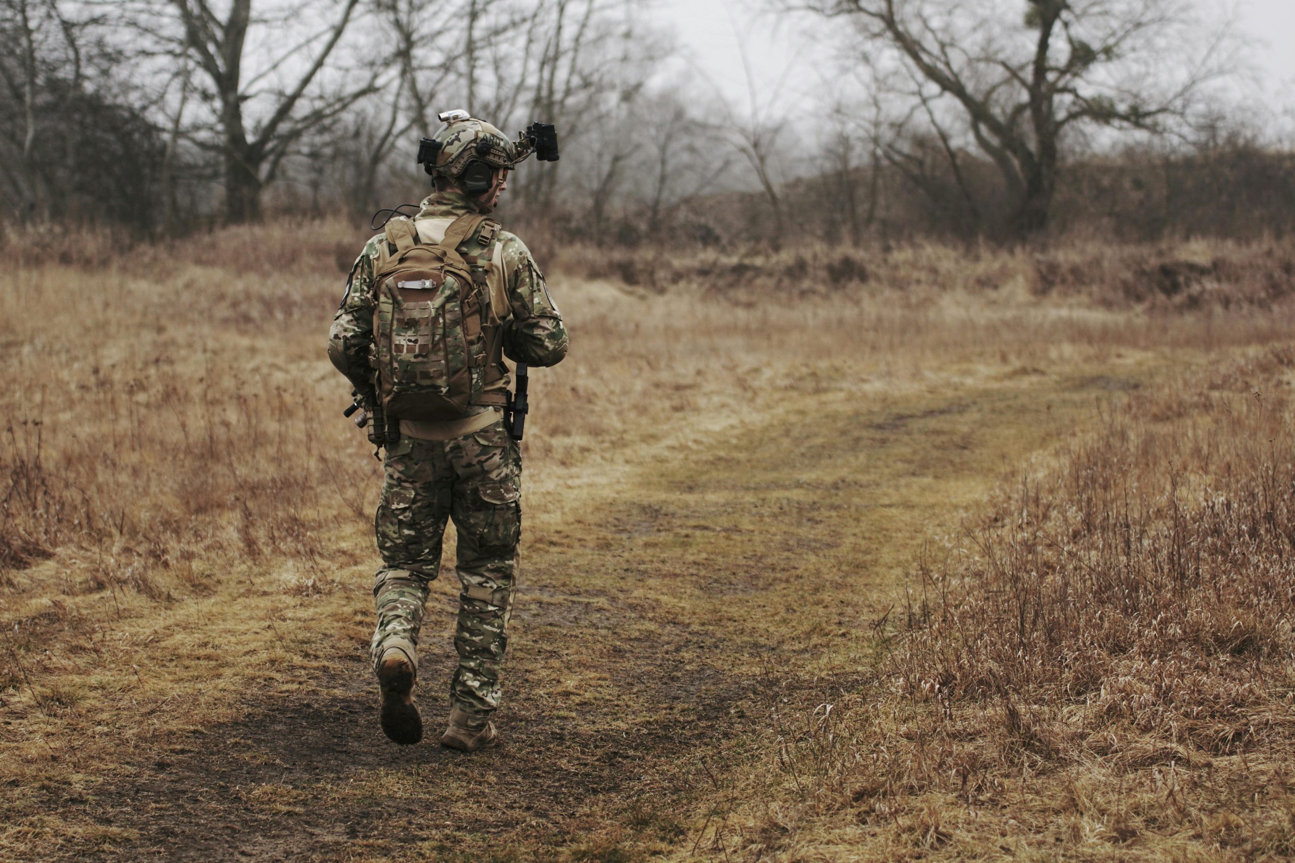 A soldier in camouflage walking along a grassy path through a forested area.