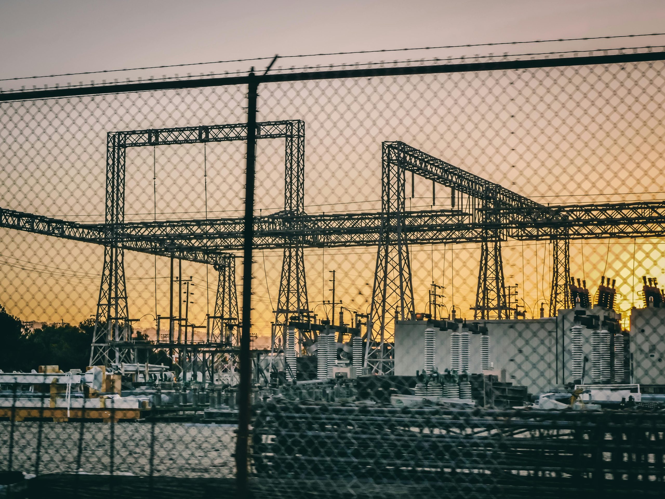 Industrial electric power substation photographed at sunset with fence in the foreground.